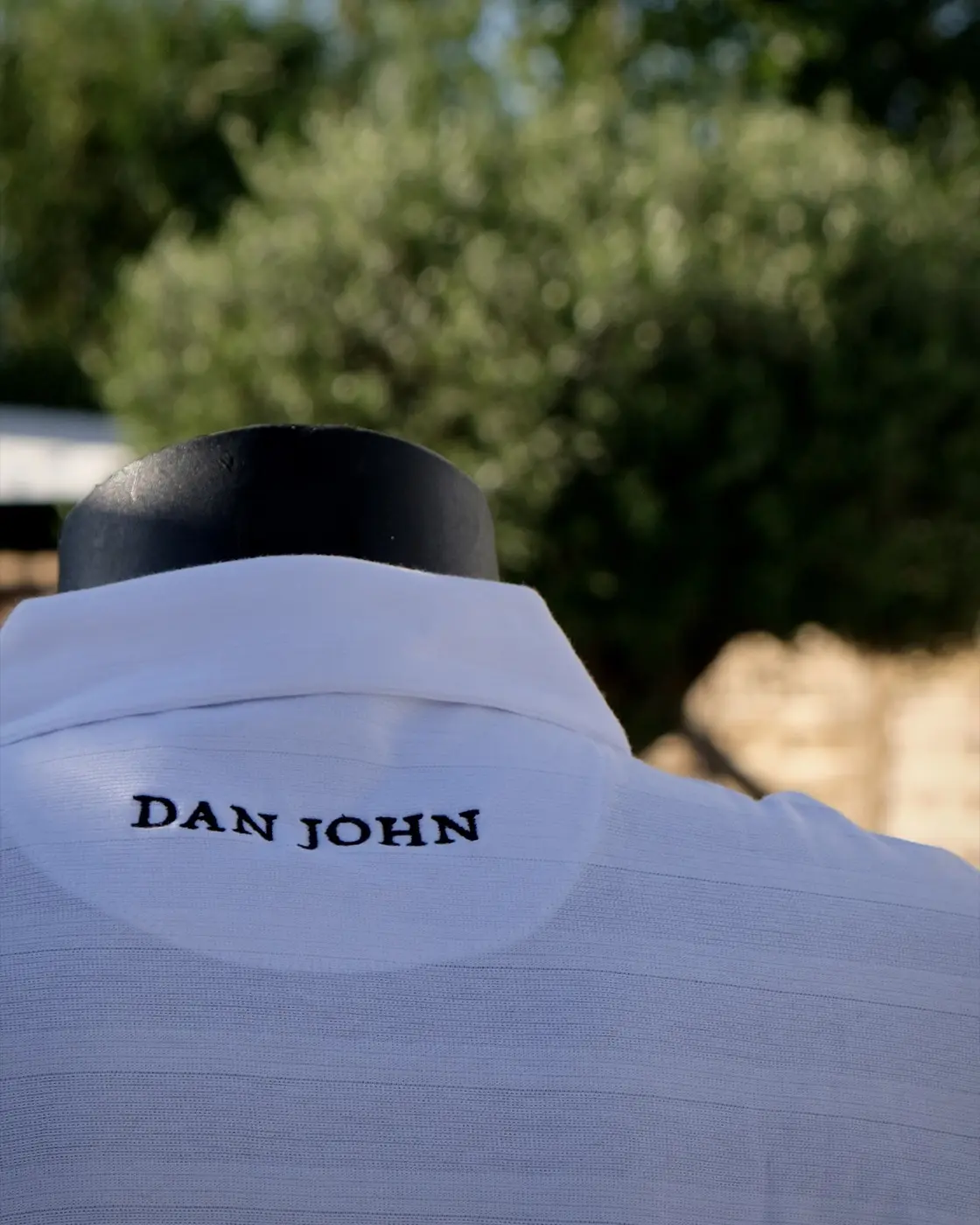 Divisa della sala del Cortile firmata Dan John: polo bianca con ricamo, sullo sfondo il verde del giardino