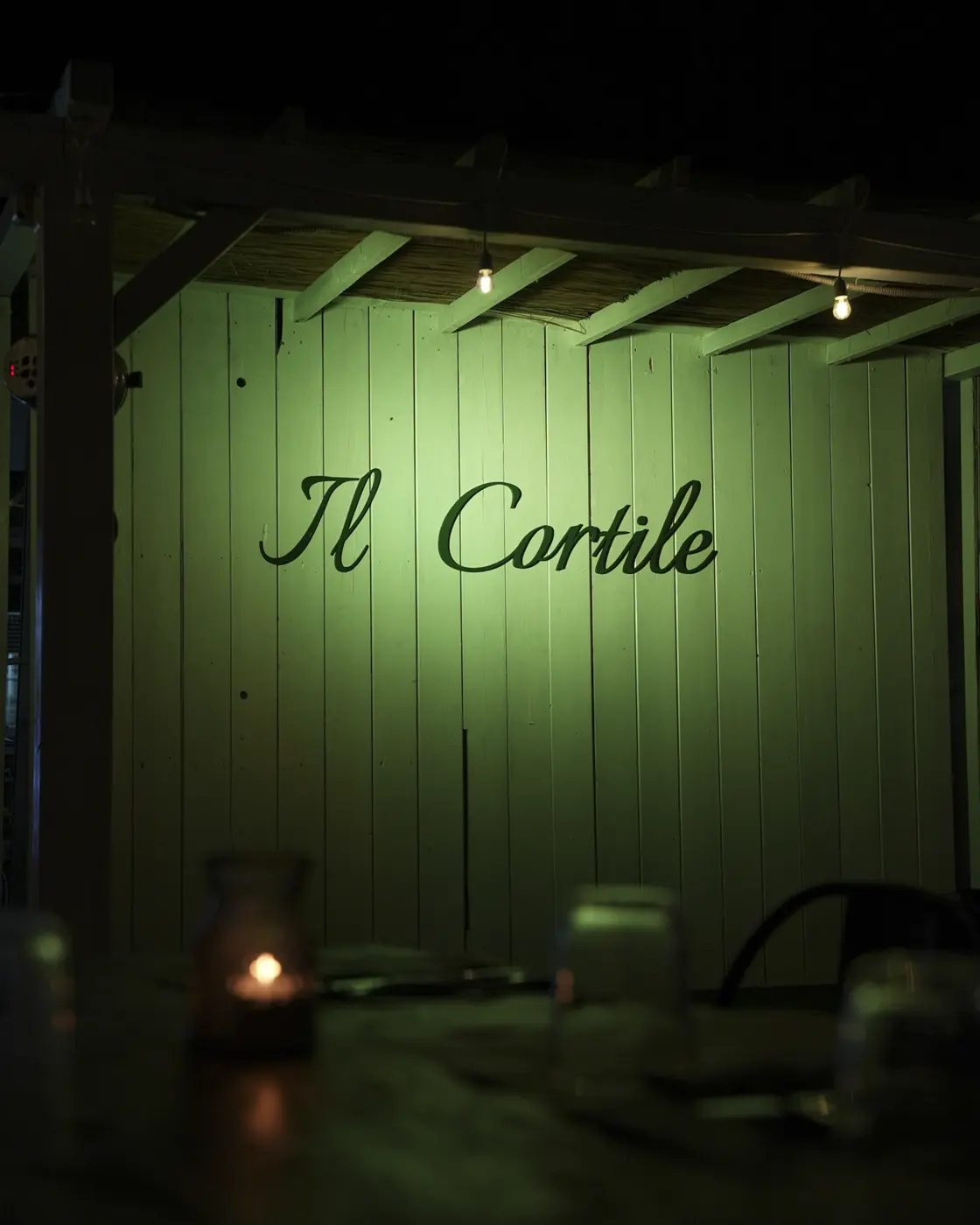 L'insegna del Cortile sulla parete verde del giardino, illuminata di sera sopra i tavoli con le candele